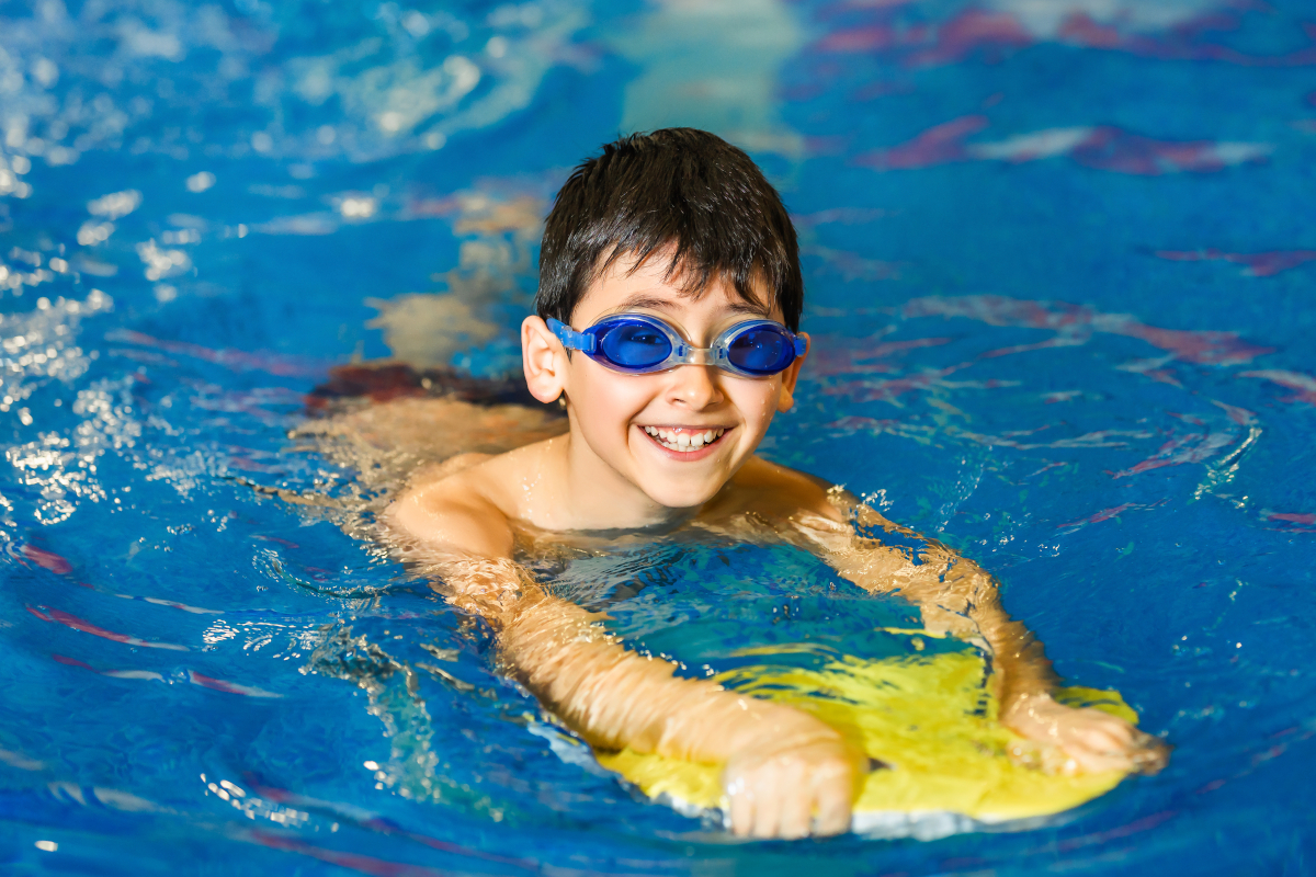 A little boy of 10 years learns to swim in the sport pool using a board. The boy wore swimming goggles. The child is cheerful and happy. Healthy kid enjoying active lifestyle.
